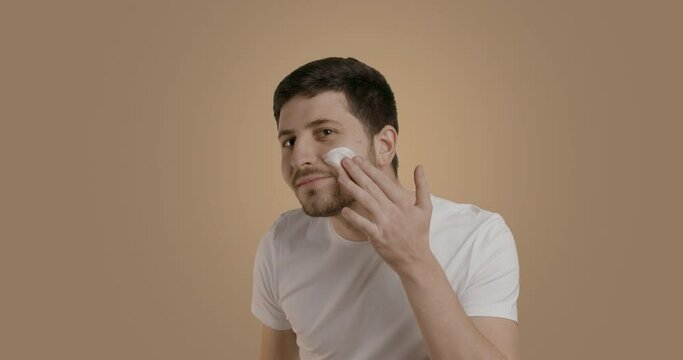 Slow motion shot of a bearded man applying a face moisturizer or face serum with two fingers. Man smiling nicely at the camera. Studio image, on light background. Concept of professional care products