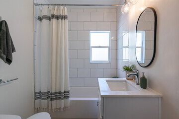 Bathroom detail of large tiled tub with shower curtain, small vanity, and black rim mirror.