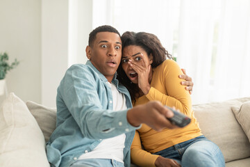 Obraz premium Scared shocked young african american woman and man with remote control switch channel on tv, watch horror film