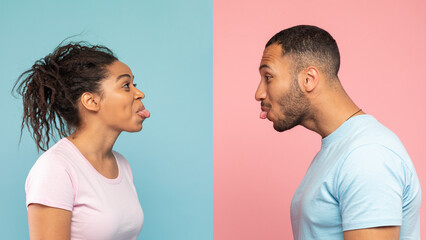 Funny black couple having fun and sticking tongue out at each other, standing over blue and pink studio background