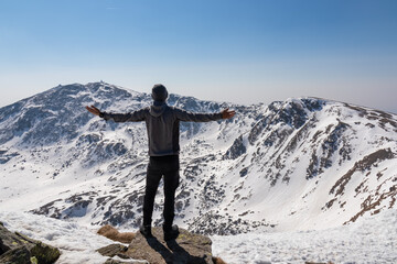Rear view of sport man on top of summit Kreiskogel spreading arms and looking at snowcapped mountain peak Zirbitzkogel, Seetal Alps, Styria, Austria, Europe. Hiking trail Central Alps in sunny winter
