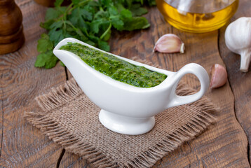 Green, spicy Cilantro Sauce with garlic and olive oil served in white ceramic sauce boat with ingredients. Selective focus, horizontal, wooden table.