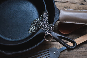 Cast iron skillets, with a metal spatula, leather handle sleeves and a chainmail scrubber on a wooden background. Cast iron cleaning and care.