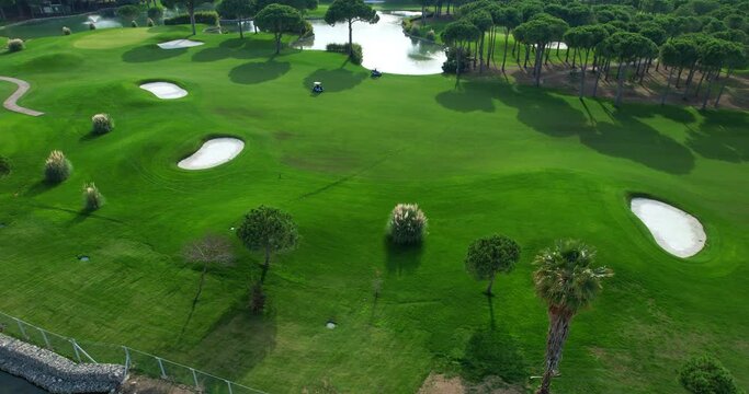 Aerial view around beautiful pond located on green golf course and tall trees casting an elegant shadow on a sunny summer day, and moving golf cars. Drone view of the clubs large golf course