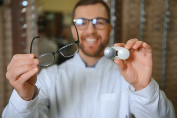 Male doctor holding contact lens case and glasses, indoors - obrazy, fototapety, plakaty