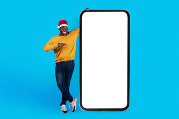 Online Offer. Happy Black Man In Santa Hat Pointing At Blank Smartphone