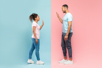Side view of young black man and woman waving to each other, standing on halved pink and blue background