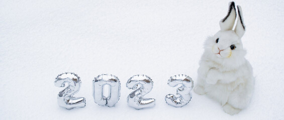 Banner with white fluffy bunny toy with silver numbers of the New Year 2023 on the white snow...