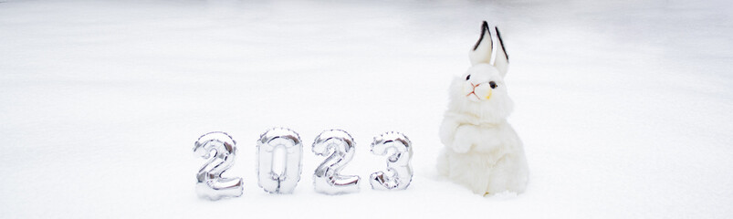 Banner with white fluffy bunny toy with silver numbers of the New Year 2023 on the white snow background. Copy space. Symbol of Chinese New Year 2023