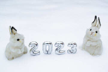 Two white cute and fluffy bunny toys with silver numbers 2023 on the white snow forest background. Copy space. Symbol of Chinese New Year 2023