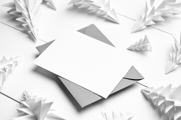 Holiday card mockup with envelope and white paper fir trees on white wooden background. Christmas greeting card mockup with decorations