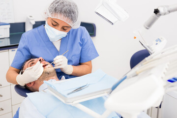 Doctor dentist woman in face mask examining a male patient teeth with dental tools