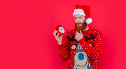 Puzzled Santa man in Christmas wear stroking beard looking at alarm clock. Christmas time