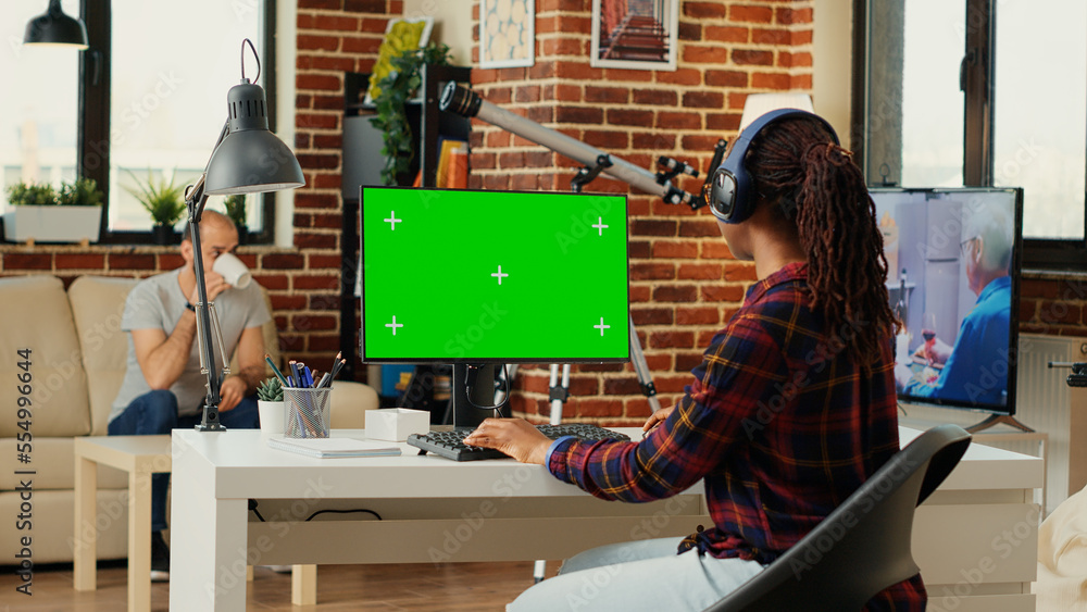 Poster office employee wearing headphones and using greenscreen on computer desktop, sitting at desk and wo