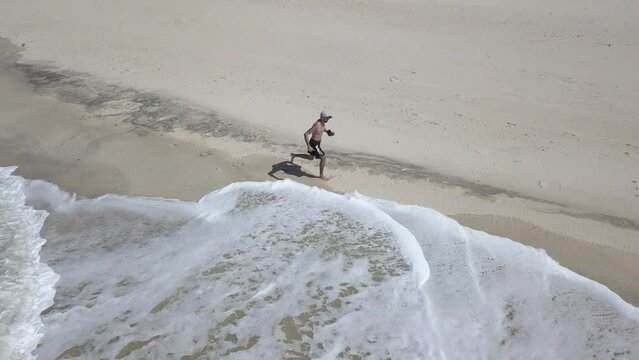 Homem sem camiseta, de bermuda e bon&eacute; correndo correndo na praia na beira do mar, segurando um controlo do drone.
