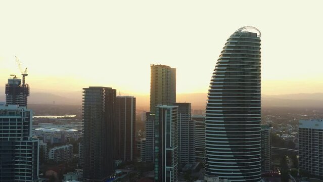 Cinematic aerial fly around waterfront apartments and resort hotels capturing broadbeach cityscape with golden glowing sun peek through in between buildings at sunset, Gold coast, Queensland.