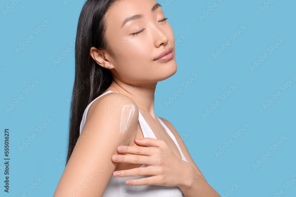 Canvas Prints Beautiful young Asian woman applying body cream onto shoulder on light blue background, space for text