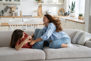 Positive young mother playing with teen girl daughter at home, carefree playful mom and child doing fun things together, mommy tickling kid while resting on sofa on weekend. Happy family concept - obrazy, fototapety, plakaty