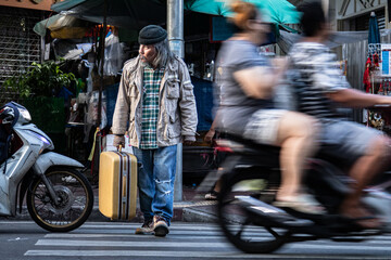 Obraz premium Asian poverty homeless man hold dragging large suitcase walk across the crosswalk in city. poor man live in slump no job, pity aged person problem from financial crisis unemployment without family