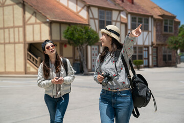 smiling asian japanese woman tourist telling sister to look at something fun in distance with pointing gesture as they are traveling in sunny solvang with Danish rural houses at background