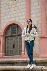 full length of leisure asian korean girl visitor wearing cap and sunglasses looking at distant view from stone steps outside old mission santa Barbara with mission door at background