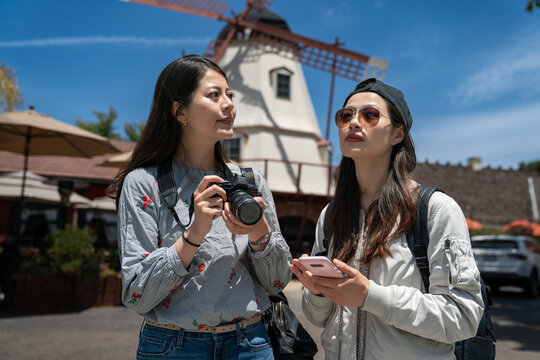 two asian korean girl traveler looking at opposite directions with windmill at background in solvang California. one is photographing famous attractions and the other is using navigation app on phone