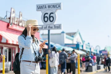 Rolgordijnen stylish asian Korean lady photographer on vacation holding hat and looking at guide on smartphone on background of route 66 end of trail sign at santa monica pier © PR Image Factory
