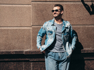 Obraz premium Portrait of handsome confident stylish hipster model. Sexy man dressed in jeans jacket. Fashion male posing in the street. In Europe city. Outdoors at sunny day. In sunglasses