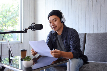 Asian man recording a podcast with headphones, microphone and script at home studio