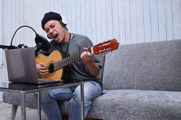 Asian guy playing the guitar recording audio podcast in studio using microphone and laptop. People and music concept.