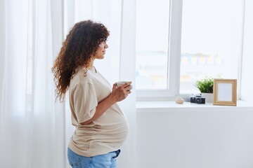 A pregnant woman stands by the window with a mug of warm water and tea and enjoys the view in a homemade t-shirt
