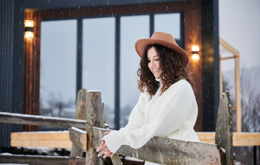 Curly young woman resting near modern barn house in the mountains. Happy female tourist enjoying winter holidays in new cottage.