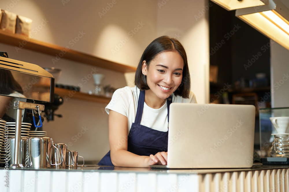 Wall mural portrait of smiling asian barista, cafe owner entrepreneur, working on laptop, processing orders on 