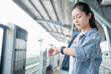 Asian woman dressing casual and looking on her watch. She standing on platform waiting for sky...
