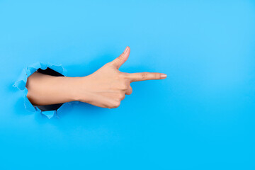 Human hand breaking through paper wall and pointing aside on virtual object or text with index finger on a blue background, space for text.