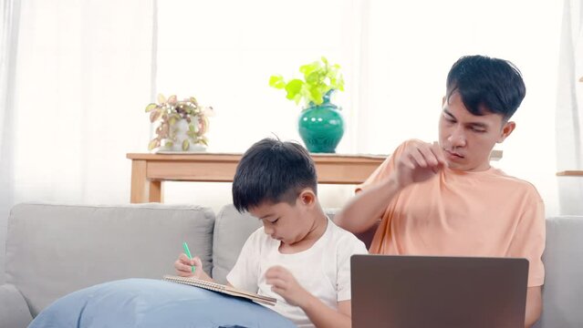 4K, An Asian single father sitting with his son on sofa living room of house, son paints picture teacher ordered, and father was working in laptop turned to kiss his son's head to cheer him up.