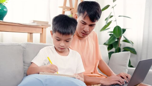 4K, An Asian single father is sitting with his son on the sofa in living room of house, teaching son to do homework to send to teacher At that time, father was working in his own notebook.