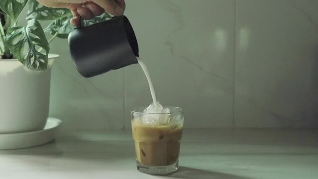 Pouring milk makes iced latte coffee into a transparent glass with ice on a white marble bar at kitchen home background movement video 4K.