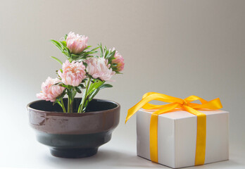 Fresh pink flowers in vase with gift box and yellow ribbon. Romantic floral arrangement, neutral background.