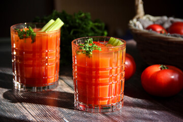 Bloody Mary cocktail with ice cubes, celery sticks and parsley leaves. Tomato juice. Red tomato cocktail in transparent glasses on a wooden table on a sunny day with tomatoes on the table. Close up.
