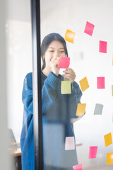 Portrait shot of Cheerful young adult business Asian woman sticking adhesive paper notes to a glass wall, idea-sharing board in business office concept.
