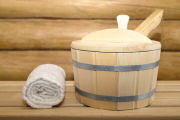 Fototapeta na wymiar A wooden bucket and towel for the spa and sauna treatments are on a wooden bench on the background log wall in sauna. 