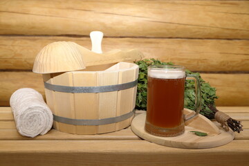 A glass mug of light beer are on a bench near the wooden bucket, ladle, birch broom and towel for the bath, spa treatments on the background of log wall in sauna.	