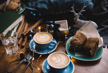 Fototapeta na wymiar Two delicious cups of cappuccino coffee standing on the rustic wooden table among Christmas decorations, fireflies, wrapped gift, scissors, cinnamon sticks, deer toy, with sandwiches on the background