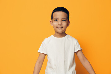 a cute, pleasant, smiling boy happily stands in a white cotton T-shirt on an orange background with an empty space for an advertising layout and looks pleasantly at the camera smiling.