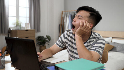 bored asian adult student propping face is playing with pen and looking away into distance with a yawn while learning from home on the computer in bedroom