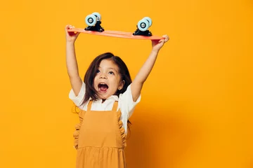 Fotobehang a joyful, happy little girl in an orange sundress stands wide open with happiness, holding a small skateboard in her hands above her head. Themes of hobbies, entertainment and outdoor activities © Tatiana