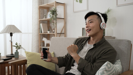 happy asian man with headphones is moving his body to the rhythm on the couch after choosing and playing songs on the smartphone at a bright home interior.