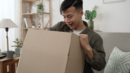 cheerful asian young male is looking into box at his online shop order on the sofa at home.
