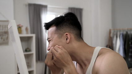 Naklejka na ściany i meble closeup with side view upset asian young guy worried about facial skin problem is trying to get rid of acnes in front of mirror in the morning at his bed chamber